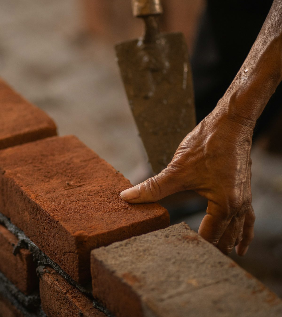 Een hand legt een rode baksteen op een muur in opbouw, met de troffel ernaast